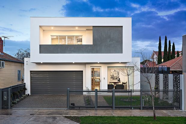 Street-facing exterior view of a contemporary two-storey residence with a crisp white rendered facade, dark grey upper-level feature panel, recessed balcony or window opening, integrated double garage, and a fenced front garden. The home is set among established suburban houses and photographed at dusk.