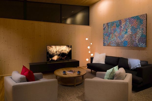 A warm, intimate living room with plywood-lined walls and ceiling, a dark built-in media console, television, low round timber coffee table, and a dark sectional sofa paired with two light upholstered armchairs. An abstract artwork provides a colourful focal point, while a sculptural cluster of globe lights adds ambient illumination. High-level dark glazing or screening is visible above the partition wall.