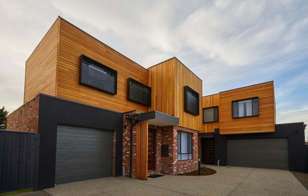 Exterior view of a contemporary multi-level residence with strong geometric volumes, warm timber cladding, exposed red brickwork, black garage doors and trim, and projecting box-like upper floors. The home features a pitched central roof form, dark-framed windows with external louvres, two integrated garages, and a textured concrete driveway.