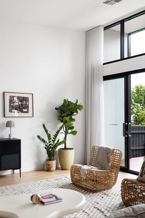 Bright contemporary living room with white walls, pale timber flooring, a textured neutral rug, woven rattan lounge chairs, potted plants, and a rounded white coffee table. Large black-framed glazed doors and upper windows open to an outdoor deck, creating an airy indoor-outdoor connection. A slim black console, framed artwork, and sheer full-height curtains complete the room.