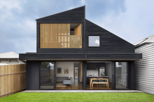 Exterior view of a contemporary two-storey residence with a dramatic asymmetrical pitched roof and dark horizontal cladding. The ground floor has extensive full-height glazing opening toward a lawn, revealing a dining area and living space. The upper facade features a warm timber vertical screening element and a projecting square window, with neighbouring homes visible on either side.