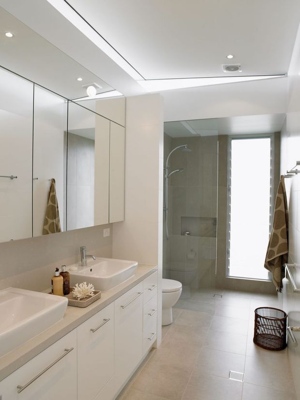 Bright, contemporary bathroom with a long white double vanity, integrated mirrored wall cabinetry, large-format light grey floor and wall tiles, and a glass-enclosed shower. A toilet, tall frosted window, heated towel rail, recessed lighting, and minimal chrome fixtures complete the clean, functional space.