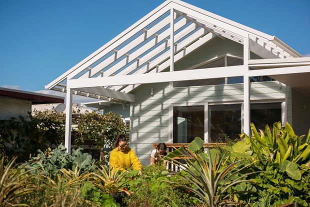 Exterior view of a light-coloured, single-storey house with a prominent white exposed gable roof structure, broad covered outdoor framing, horizontal weatherboard-style cladding, and large glazed openings. Lush subtropical planting fills the foreground, with people seated near the covered outdoor area. The image is captured in bright daylight against a clear blue sky.