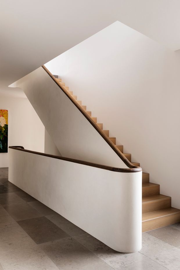 A minimalist interior stairway with a sculptural curved plaster balustrade and dark timber handrail. Timber treads rise alongside a smooth white wall, while large-format stone or concrete-look flooring extends across the lower level. The composition is softly lit with neutral, refined finishes and a glimpse of artwork at the far left.