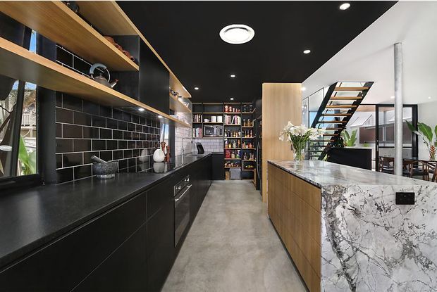 A contemporary galley-style kitchen with black cabinetry, open timber shelving, and a black subway-tile splashback. A large engineered-stone island with dramatic grey veining occupies the foreground. The space has a polished concrete floor, black ceiling with recessed lighting, timber accents, and an open connection to a dining or living area with a floating staircase.