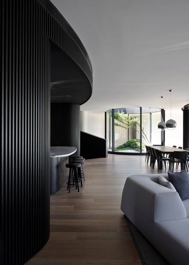 A refined open-plan interior combining a living room, dining area and compact bar. The space features dramatic curved black ribbed wall and ceiling elements, dark-framed curved glazing around a planted courtyard, warm timber flooring, a pale sectional sofa, and a long dark dining table beneath sculptural spherical pendant lights. The palette is restrained and contemporary, with strong contrast between black architectural features, light walls and warm wood.