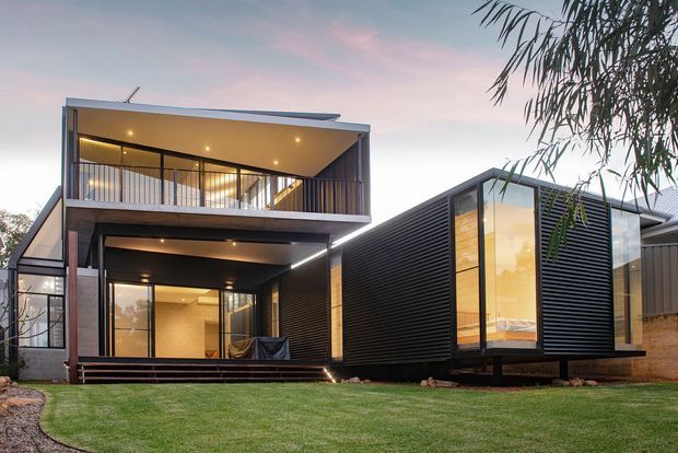 Contemporary multi-level residential building viewed from the lawn at dusk. The home features strongly articulated rectangular volumes, a projecting upper balcony, extensive floor-to-ceiling glazing, dark horizontal cladding, exposed structural elements, and warm interior lighting. A smaller dark-clad wing with a tall glazed end sits beside the main volume.