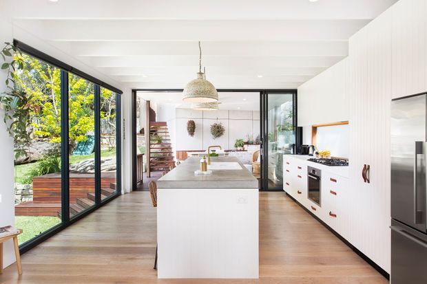 Bright contemporary kitchen with a large white island, white fixed cabinetry, integrated appliances, timber flooring, and black-framed floor-to-ceiling glazing opening to a leafy garden. A sculptural pendant hangs above the island, while a dining and living area with open timber stairs is visible beyond.