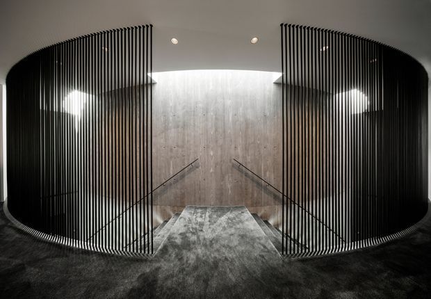 Dramatic contemporary stairway or landing with a symmetrical curved black vertical-slat screen enclosing the sides. A central concrete-textured feature wall forms the backdrop, with slim dark handrails descending on either side and dark plush carpet covering the floor and steps. Recessed ceiling lights and concealed illumination create a refined, monochromatic interior.