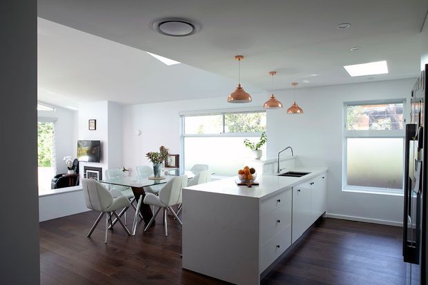 Bright open-plan kitchen and dining area with a large white island, integrated sink and cooktop, white cabinetry, dark timber flooring, copper pendant lights, skylights, and ample windows. A glass dining table with upholstered chairs sits beside the kitchen, with a small sitting area visible in the background.