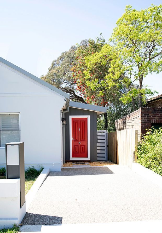 Bright exterior view of a compact residential entry. A vivid red paneled front door with white trim is set into a dark grey vertical-clad entry extension beside a white-painted main facade. The approach is a light aggregate or concrete driveway/path, bordered by a low white wall and a timber fence, with a brick boundary wall and mature trees beyond.