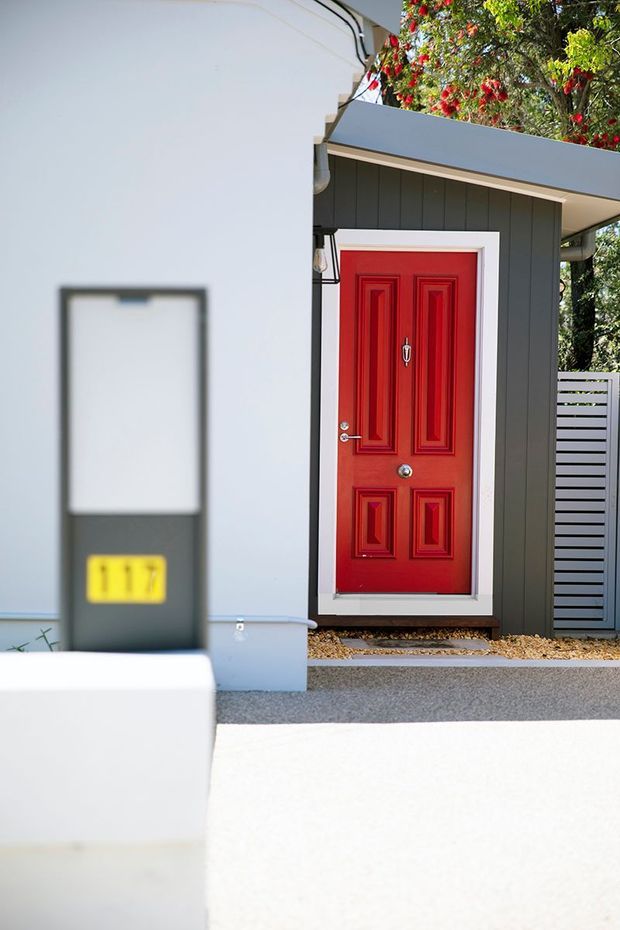 Close-up exterior view of a contemporary residential entry. A vivid red paneled front door with white trim is set within a dark charcoal vertical-clad porch structure beneath a projecting skillion-style canopy. The foreground includes a blurred mailbox and number plaque, while a horizontal slatted privacy fence and leafy planting appear to the side and above.