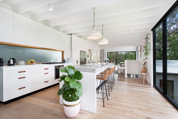 Bright open-plan kitchen and living/dining area with white flat-panel cabinetry, a large central island, timber flooring, black-framed floor-to-ceiling glazing, and exposed white ceiling beams. Woven bar stools and pendant lights add warmth to the clean contemporary space, with greenery visible through the glazing.
