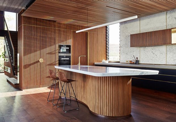 A sophisticated open-plan kitchen with a large curved island, integrated appliances, dark timber flooring, and extensive vertical timber slat joinery. The island has a pale stone benchtop with a waterfall edge and seating for two. A textured light stone or concrete-look backsplash contrasts with the warm timber walls and slatted ceiling. Black cabinetry, linear pendant lighting, and a glimpse of a stairway contribute to the refined contemporary design.