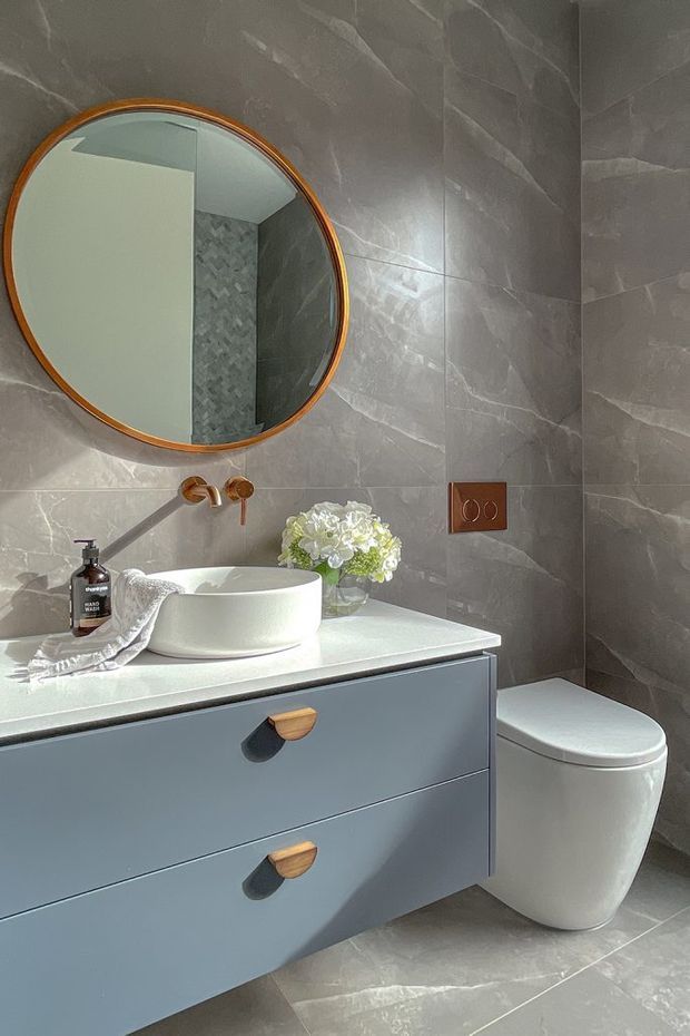 A compact, refined bathroom featuring large grey marble-effect wall and floor tiles, a wall-mounted blue-grey vanity with timber and dark inset pulls, a white vessel basin, and a modern white toilet. A round mirror with a warm timber frame hangs above the vanity, alongside copper-toned tapware and flush controls. Soft daylight and a small floral arrangement add warmth to the otherwise cool-toned space.