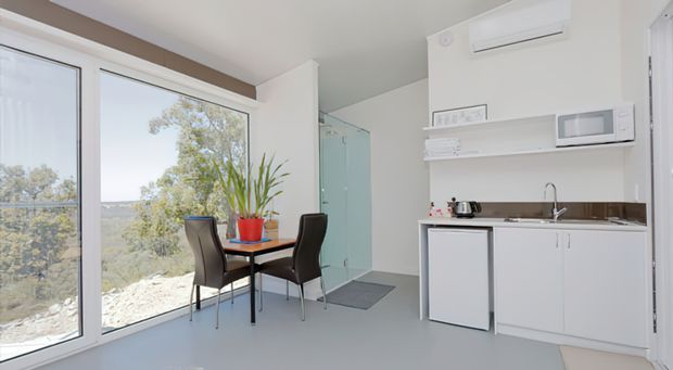 Bright compact open-plan interior with a small dining table and four chairs beside full-height glazing, overlooking a natural landscape. A minimalist white kitchenette with integrated under-counter refrigerator, sink, microwave, open shelving, and wall-mounted air conditioner occupies the right side. A glass shower enclosure is visible behind the dining area, suggesting a compact studio or apartment layout.
