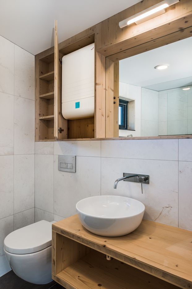 Compact contemporary bathroom with large pale stone-look wall tiles, a white vessel basin on a built-in timber vanity, wall-mounted chrome tapware, concealed cistern toilet, and a mirrored timber-framed recessed cabinet with open shelving and integrated lighting.
