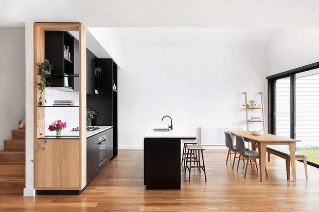 Bright open-plan kitchen and dining area with warm timber flooring, a compact black kitchen island, tall black and timber cabinetry, a timber dining table with upholstered chairs, and large black-framed glazing. A timber staircase and built-in shelving are visible at left, with minimalist styling throughout.
