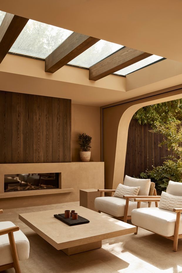 Warm, refined living room with a contemporary architectural interior. The space features a large central stone or plaster-finished coffee table, cream upholstered timber-framed lounge chairs, a built-in fireplace with dark vertical timber panelling, and a curved plaster arch opening to a planted courtyard. Exposed dark timber beams support multiple skylights, creating strong natural light and shadows. The palette is earthy and neutral, with beige plaster, natural wood, cream upholstery, and dark brown accents.