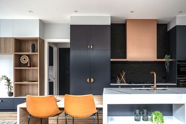 Contemporary kitchen featuring dark cabinetry, warm timber open shelving, a black veined splashback, copper-toned rangehood and tapware, and a pale stone island with open display shelving. Two orange counter stools add a vivid accent, while integrated appliances and recessed ceiling lights create a clean, tailored fit-out.