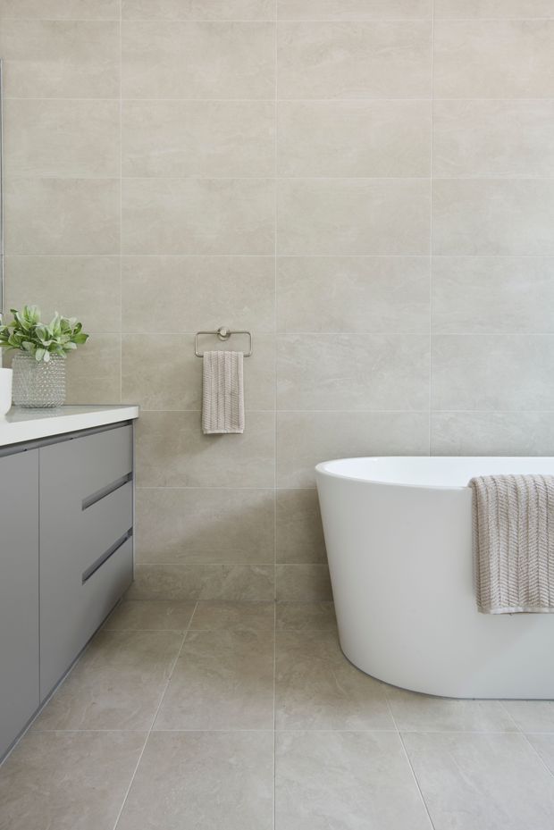 A contemporary bathroom with large-format light beige-grey stone-look tiles covering the walls and floor. The room features a freestanding white bathtub, a minimalist grey vanity with slim integrated pulls and a pale benchtop, a neutral textured hand towel, and a small potted plant. The overall design is clean, calm, and understated.