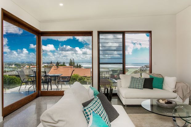 Bright coastal living room with white sofas, a glass coffee table, and expansive timber-framed glazing overlooking rooftops, ocean, and sky. The room has a clean, relaxed feel with turquoise and patterned cushions, a neutral rug, and polished light flooring.