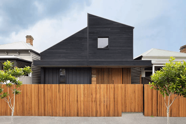 Front exterior view of a compact contemporary home with a distinctive asymmetrical gabled roof form, dark horizontal timber cladding, vertical timber screening and a warm timber privacy fence. The house is set between neighbouring traditional homes, with a small gated entry and young street trees.