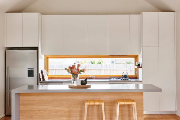 Bright, compact contemporary kitchen with a large central island, pale flat-panel cabinetry, a timber-clad island front, stainless-steel refrigerator, and a warm timber-framed horizontal window above the cooktop and benchtop. Two timber stools sit at the island, and the vaulted ceiling adds openness. The palette is restrained and warm, with white cabinetry, light timber, and neutral stone-like surfaces.