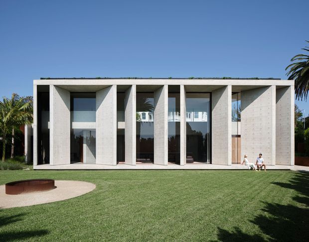 A modern, single-storey concrete house viewed from the rear garden. The rectilinear facade is composed of deep projecting and recessed concrete frames forming a regular colonnade around full-height glazed openings. The flat roof has a low planted or green edge, and the expansive lawn includes a curved paved area with a shallow dark fire bowl or water feature. Palm trees and a bright blue sky surround the property.