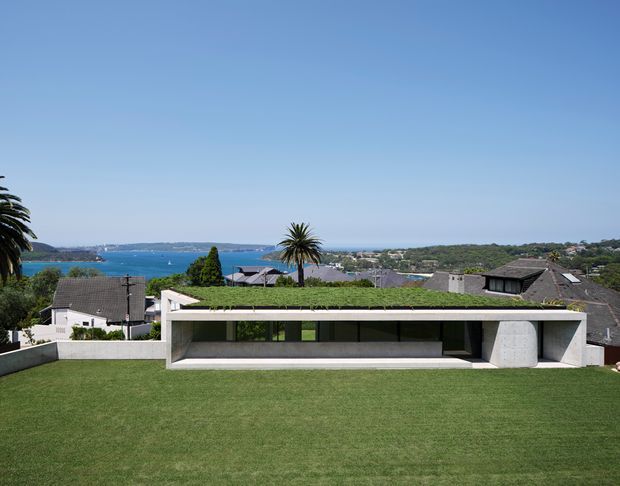 Exterior view of a low, contemporary concrete house with a broad flat roof planted with grass, a long recessed glazed opening, and a large manicured lawn in the foreground. The property overlooks a blue coastal inlet with residential surroundings and palm trees in the distance.