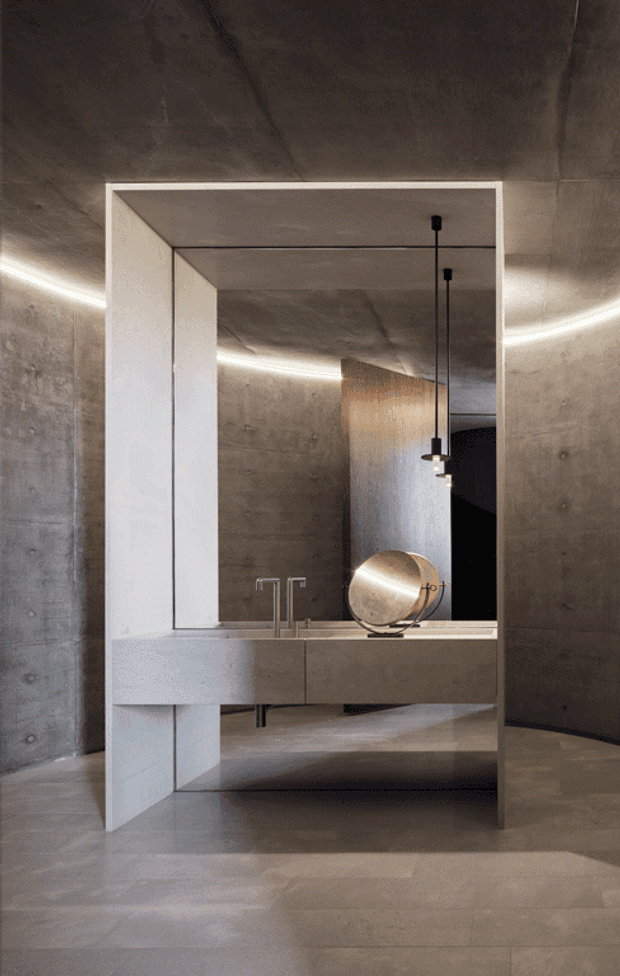 A dramatic contemporary bathroom vanity installation set within a large white-framed open cabinet or portal. The space features raw concrete walls, ceiling and floor, a pale integrated vanity with twin faucets, a round metallic basin or vessel, black pendant lighting, and concealed linear LED lighting tracing the wall and ceiling junctions. A reflective opening reveals the continuation of the concrete interior and timber-toned panel.