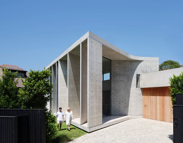Contemporary concrete residential facade with a sculptural curved roof and canopy, tall recessed glazing, projecting concrete fins, and a warm timber garage door. The house is viewed from the driveway, with landscaped planting, boundary fencing, and neighbouring homes visible around it.