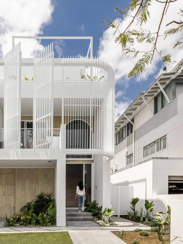 Street-facing view of a bright white multi-storey townhouse or residential facade. The building features a rounded projecting balcony enclosed by vertical white metal screening, a tall rectangular rooftop frame, arched glazing at the entry level, pale timber-look garage doors, and lush tropical planting along a concrete path. A neighbouring white building with louvred windows and exposed roof structure is visible to the right under a blue sky.