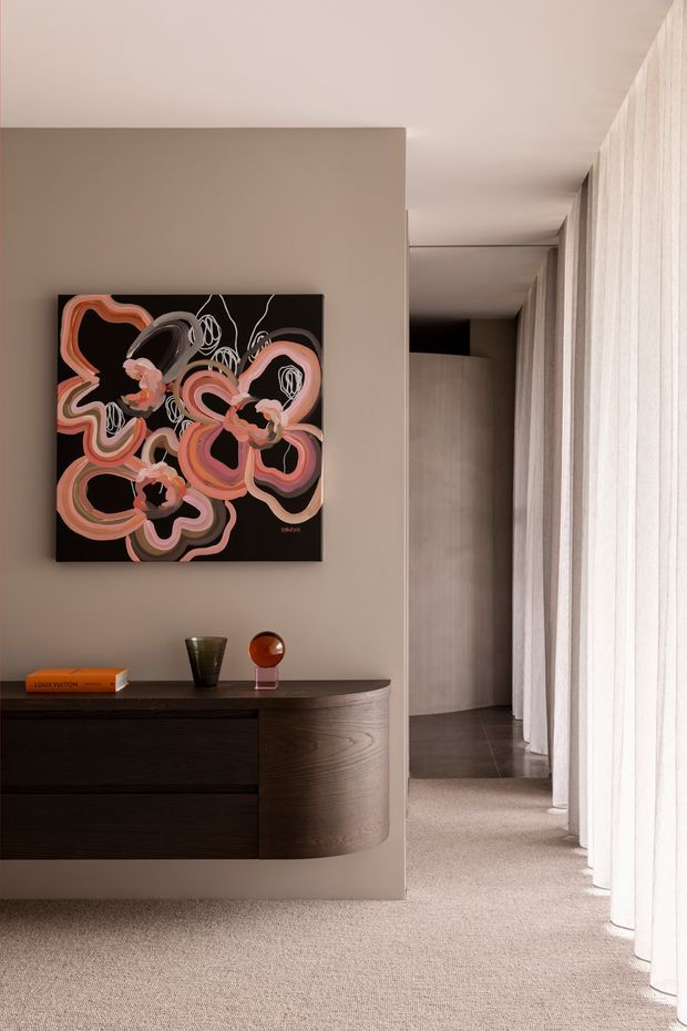 A refined residential hallway or landing with a long wall of full-height sheer curtains on the right, a warm taupe plaster wall, and a dark curved timber console with drawers. An abstract artwork in coral, pink, cream and black hangs above the console, accompanied by small decorative objects. The corridor continues toward a darker timber-lined area, while a soft neutral carpet creates a calm, upscale atmosphere.