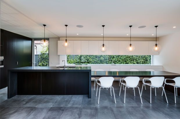 Contemporary open-plan kitchen and dining area with a large dark island, extensive pale built-in cabinetry, a long horizontal garden-facing window, pendant lighting, and a dining table surrounded by white chairs. Large-format grey floor tiles and dark timber cabinetry create a refined, high-contrast interior.