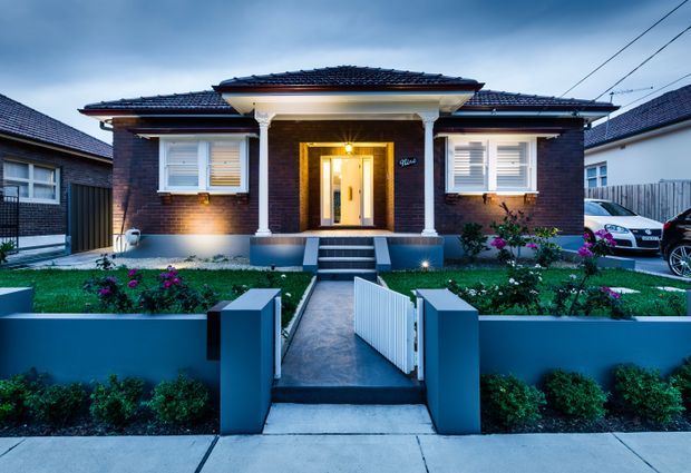 Front exterior view of a single-storey brick home with a symmetrical entrance porch, white columns, tiled hip roof, white-framed windows, landscaped front garden, low rendered boundary walls and a central pedestrian gate. The home is photographed at dusk with warm entry lighting and cool blue exterior tones.