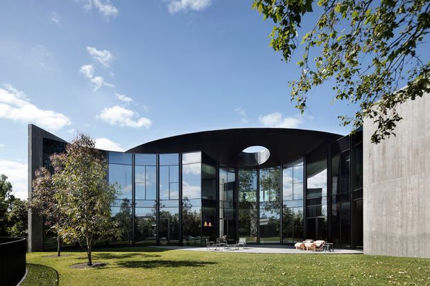 A striking contemporary building with a sweeping curved dark roof and expansive floor-to-ceiling glass walls surrounding a landscaped lawn courtyard. A circular roof opening creates a dramatic oculus above the glazed interior, while a tall raw concrete wall forms one side of the composition. Outdoor seating is visible near the glazing, with mature trees and a clear sky surrounding the property.