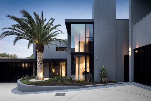 Contemporary multi-level residential facade at dusk, composed of smooth dark-grey rendered walls, black metal garage and entry gates, and extensive floor-to-ceiling glazing around a tall entry volume. A curved raised planter with a palm and low landscaping fronts the house, while patterned paving and a broad driveway lead to the entrances. Warm interior lighting contrasts with the cool exterior palette.