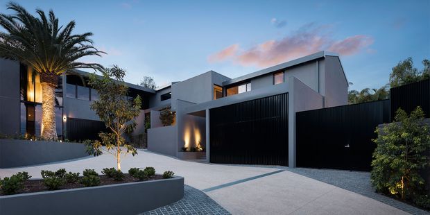 A dusk exterior view of a large contemporary residential property, featuring crisp cubic forms, dark charcoal rendered walls, extensive black vertical metal screening, a tall integrated garage door, and a broad pale paved driveway. Warm architectural lighting highlights the entry and landscaping, with mature palm trees and carefully designed raised garden beds framing the facade.