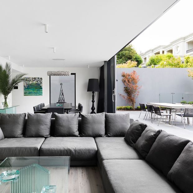 Contemporary open-plan living and dining area with a large charcoal-grey sectional sofa in the foreground, glass coffee table, dark dining table and chairs, sculptural pendant light, floor lamp, artwork, and indoor greenery. Full-height glazed doors open to a paved outdoor dining terrace with planting and a rendered boundary wall. The room has a bright, restrained neutral palette and strong indoor-outdoor connection.