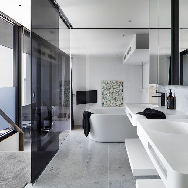 Bright contemporary bathroom with a freestanding white bathtub, double wall-mounted vanity basins, large mirror, black tapware, marble-look tiled flooring, and a black-framed glass partition. A compact living area is visible beyond the partition, including a wall-mounted television and artwork.