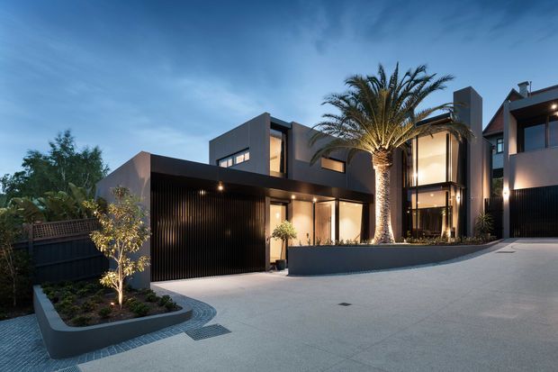 A dusk exterior view of a contemporary multi-storey residence with a dark geometric facade, large glazed openings, a broad driveway, and an integrated garage. Raised curved planter beds with tropical landscaping and a prominent palm tree soften the minimalist architecture. Warm architectural lighting illuminates the entry and upper-level glazing.