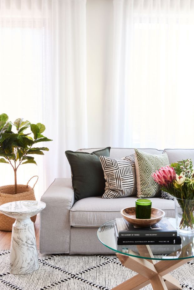 Bright, softly styled living room with a light grey upholstered sofa, sheer white floor-to-ceiling curtains, patterned monochrome and sage cushions, a glass coffee table with timber crossed-leg base, and a textured black-and-white rug. A potted indoor tree, flowers, books, and a small sculptural side table add natural and decorative detail.
