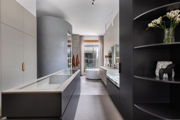 A refined contemporary bathroom with a freestanding bathtub at the rear, a long double vanity with integrated basin and pale stone surface, a large tiled cylindrical feature wall, dark built-in cabinetry and open shelving, and a partially enclosed shower or wet-area zone. The room has a restrained monochromatic palette with warm timber and brass details, soft natural light, and carefully styled decorative shelving.