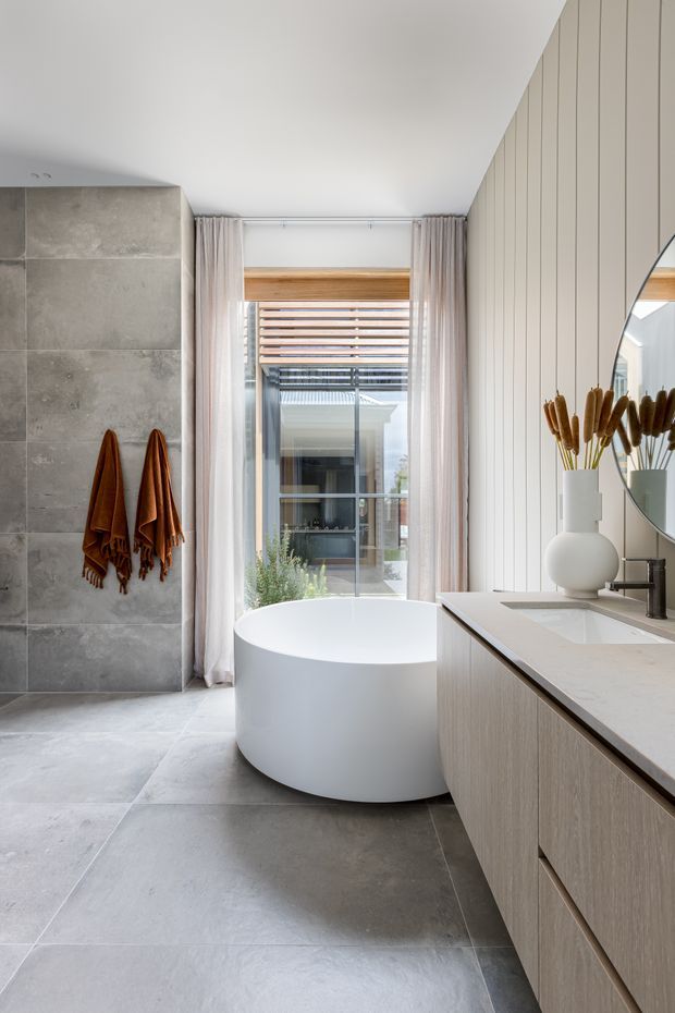 A bright, refined contemporary bathroom featuring a freestanding white oval bathtub positioned beside a large window, a floating light-wood vanity with a pale stone benchtop, a round mirror, grey large-format tiled walls and flooring, and warm timber window detailing. Soft curtains and natural light create an airy atmosphere.