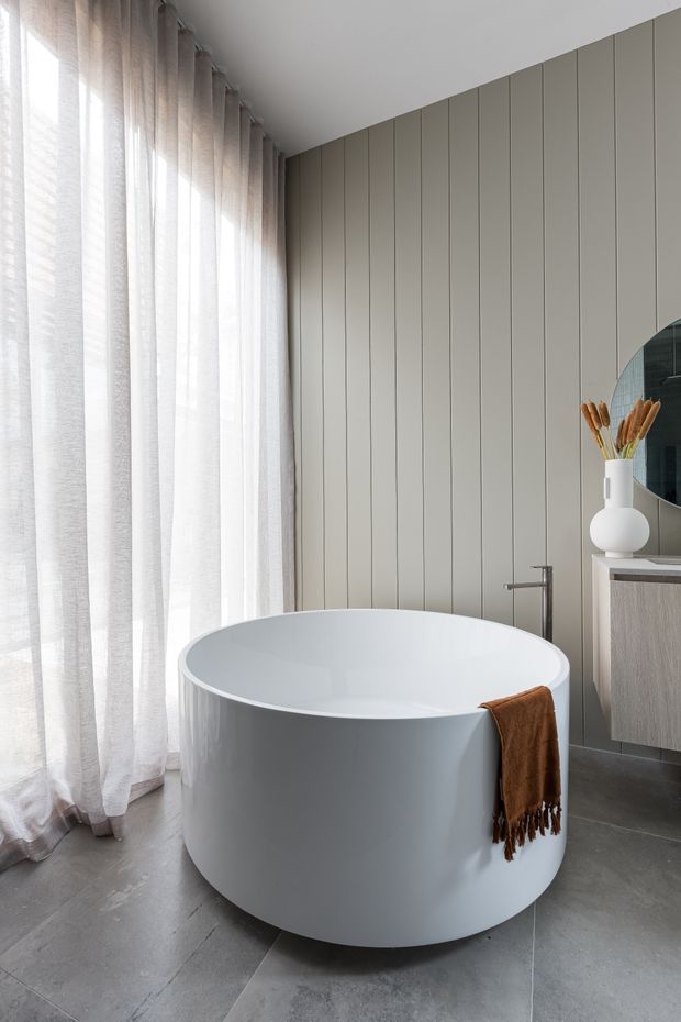 A bright, contemporary bathroom featuring a freestanding white oval bathtub, large grey floor tiles, a vertically panelled beige-grey wall, sheer full-height curtains, and a floating vanity with a round mirror. A warm brown towel adds contrast.