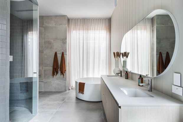 Bright contemporary bathroom with a freestanding round bathtub, walk-in glass shower, double vanity with integrated basins, large rounded mirror, pale vertical wall panelling, grey stone-look wall and floor tiles, and sheer curtains over a window. Warm brown towels and timber-toned vanity details add subtle contrast.
