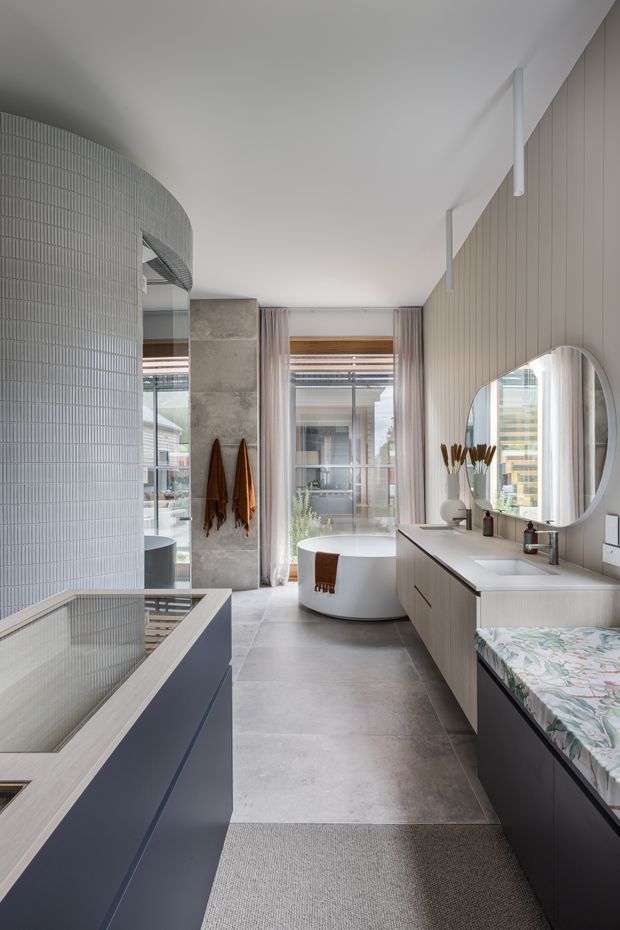 Contemporary bathroom with a freestanding oval bathtub positioned in front of full-height glazing, a large double vanity with pale wood cabinetry and round mirrors, and a deep blue tiled bathtub surround. The space features large grey floor tiles, vertically ribbed pale wall tiles, soft curtains, and restrained neutral finishes with abundant natural light.