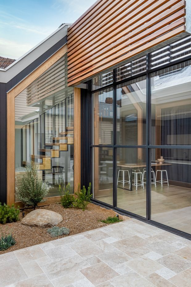 Exterior view of a contemporary residential extension opening onto a paved courtyard. The facade combines extensive black-framed glazing, warm horizontal timber screening, light timber cladding, and dark metal accents. A glazed interior reveals a kitchen or bar area and a floating timber staircase, while the foreground includes pale stone paving and a minimalist drought-tolerant garden with gravel and rocks.