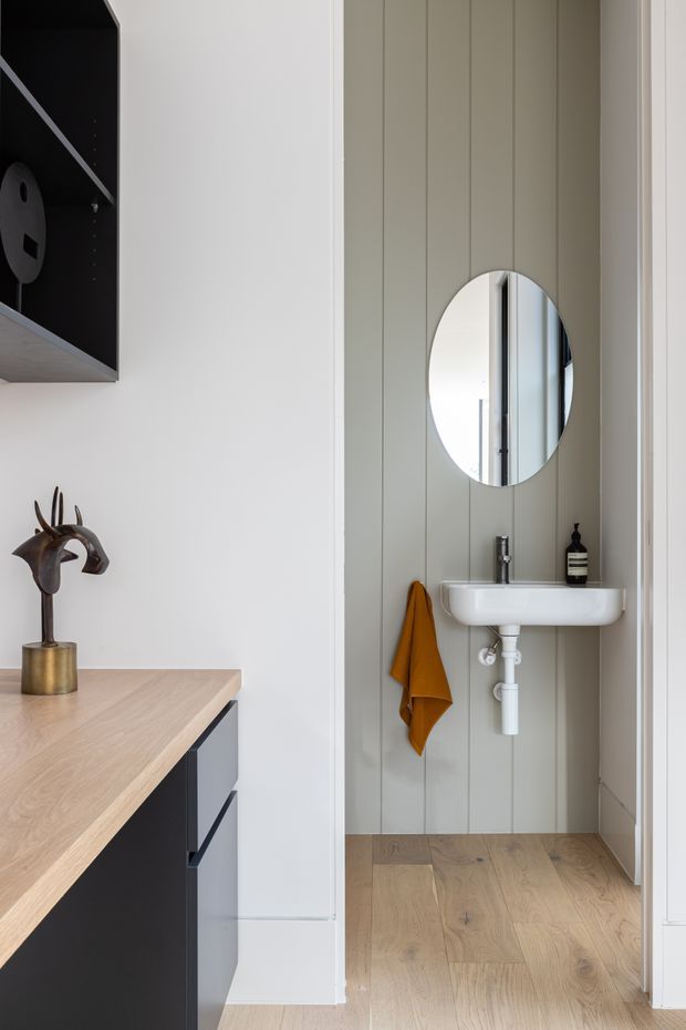 A refined compact powder room viewed through an opening, featuring a wall-mounted white basin with exposed plumbing, round mirror, vertical grey-green wall panelling, timber flooring, and a mustard towel. In the foreground is fixed dark cabinetry with a light timber benchtop and sculptural decor.